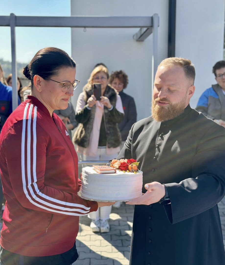 Bearded man in black clerical attire hands a white frosted cake to a woman in a red track jacket; onlookers photograph in the background outdoors.