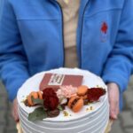 Person in a blue jacket holds a white-frosted cake decorated with orange macarons, red roses, a pink chrysanthemum, gold beads, and a chocolate plaque.