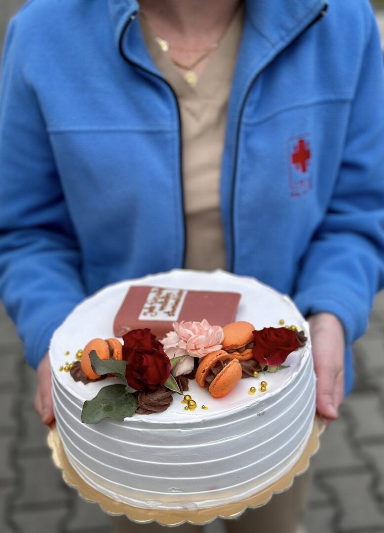 Person in a blue jacket holds a white-frosted cake decorated with orange macarons, red roses, a pink chrysanthemum, gold beads, and a chocolate plaque.