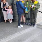 Three youths in green uniforms stand on stone steps, receiving items and flowers from a boy while an adult woman and a young girl watch from the doorway.