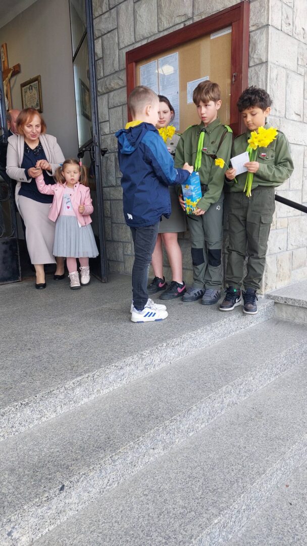 Three youths in green uniforms stand on stone steps, receiving items and flowers from a boy while an adult woman and a young girl watch from the doorway.