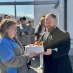 Bearded man in a black clerical robe hands a white frosted cake to an older woman in a blue-gray jacket during an outdoor celebration.