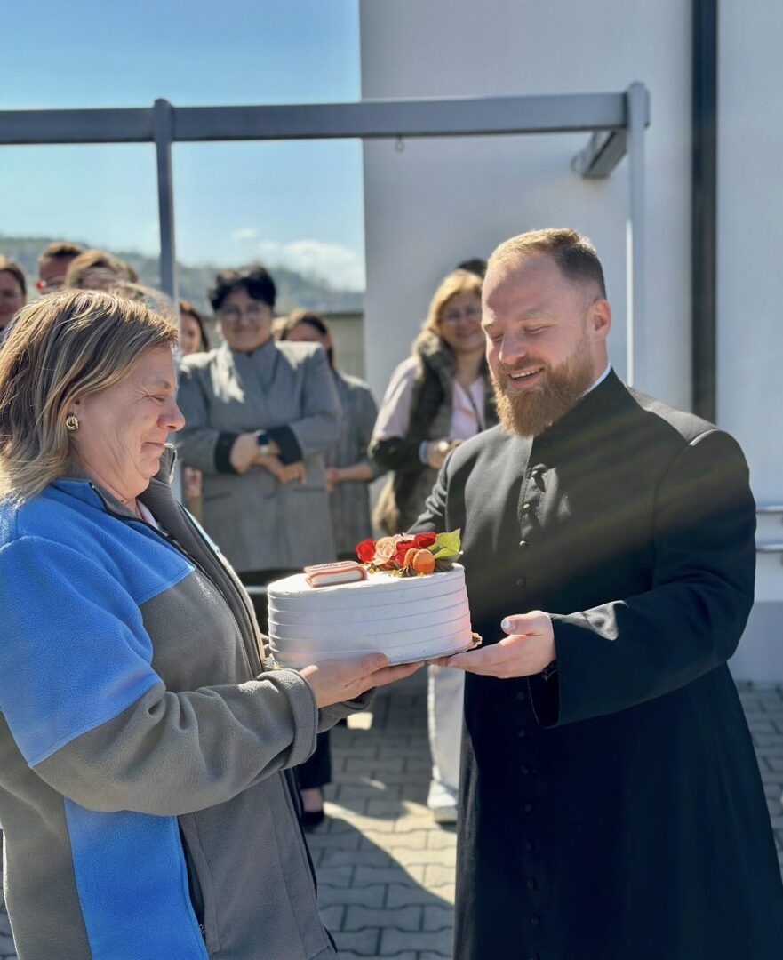 Bearded man in a black clerical robe hands a white frosted cake to an older woman in a blue-gray jacket during an outdoor celebration.