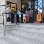 Group of children and a woman standing on stone steps outside a building entrance, looking at papers
