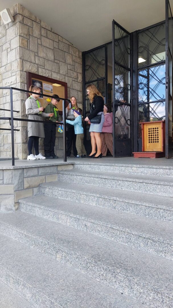 Group of children and a woman standing on stone steps outside a building entrance, looking at papers
