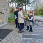 A woman giving a yellow flower bouquet to a girl in a gray uniform while other students stand nearby outside a stone building.