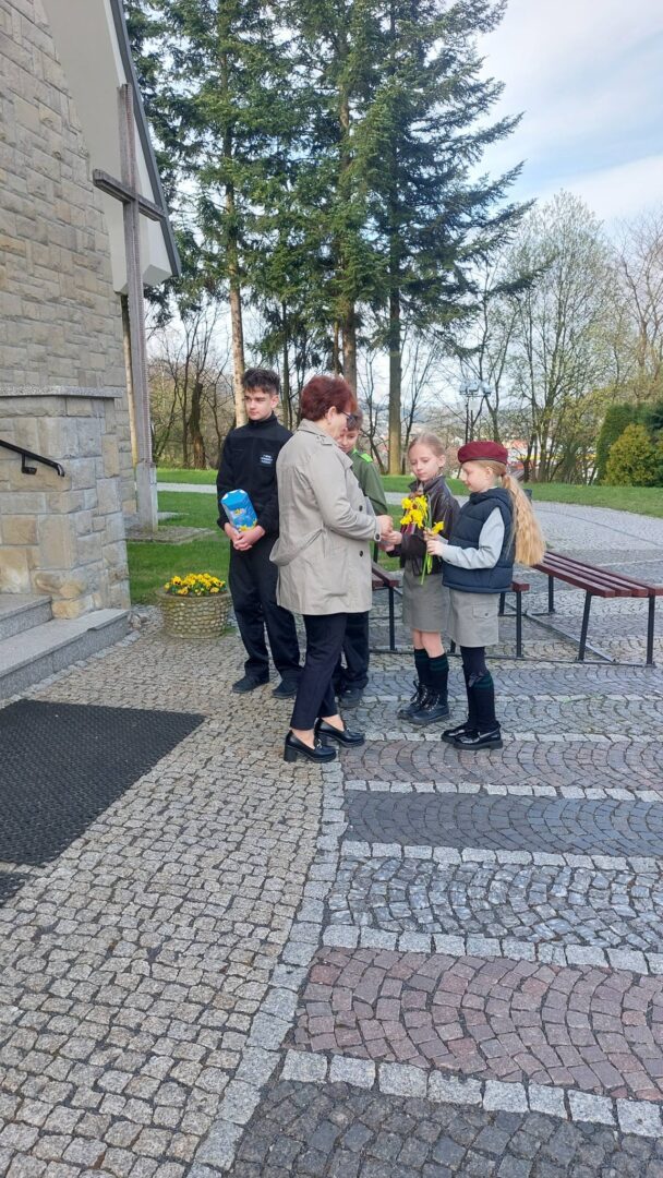 A woman giving a yellow flower bouquet to a girl in a gray uniform while other students stand nearby outside a stone building.