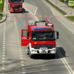 Red fire engine with blue flashing lights driving toward the camera on a city street; side door open and equipment rack visible is on top of the cab.
