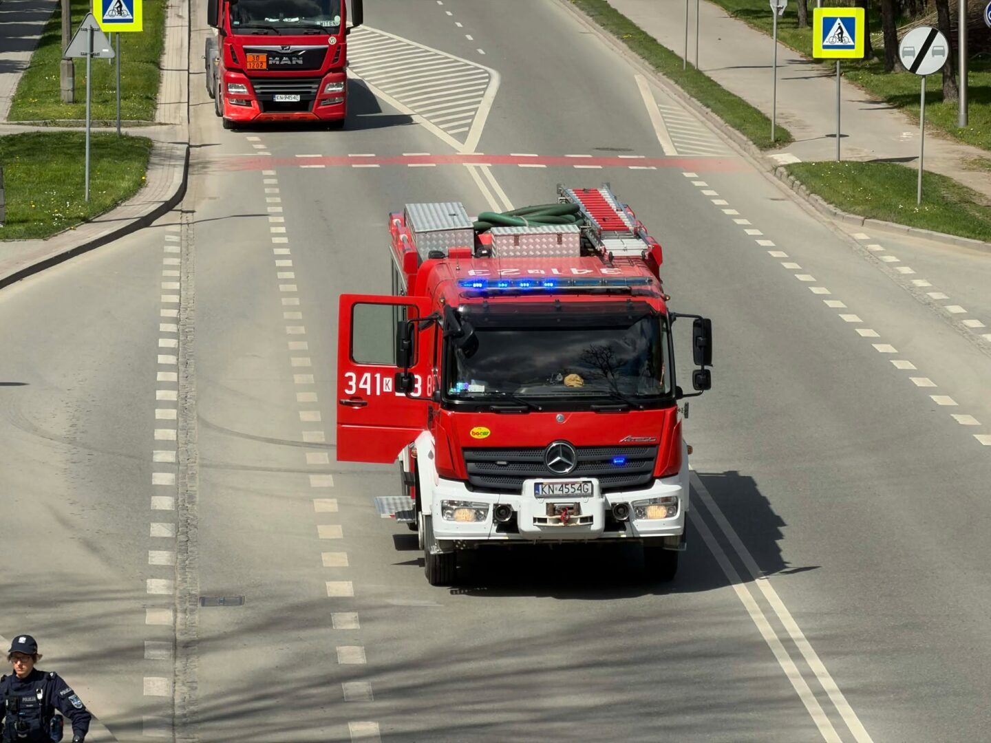 Red fire engine with blue flashing lights driving toward the camera on a city street; side door open and equipment rack visible is on top of the cab.