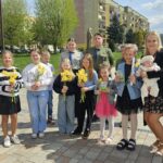 A group of children and a few adults stand outdoors in a park-like area, many holding yellow daffodils and gift cards, smiling at the camera.