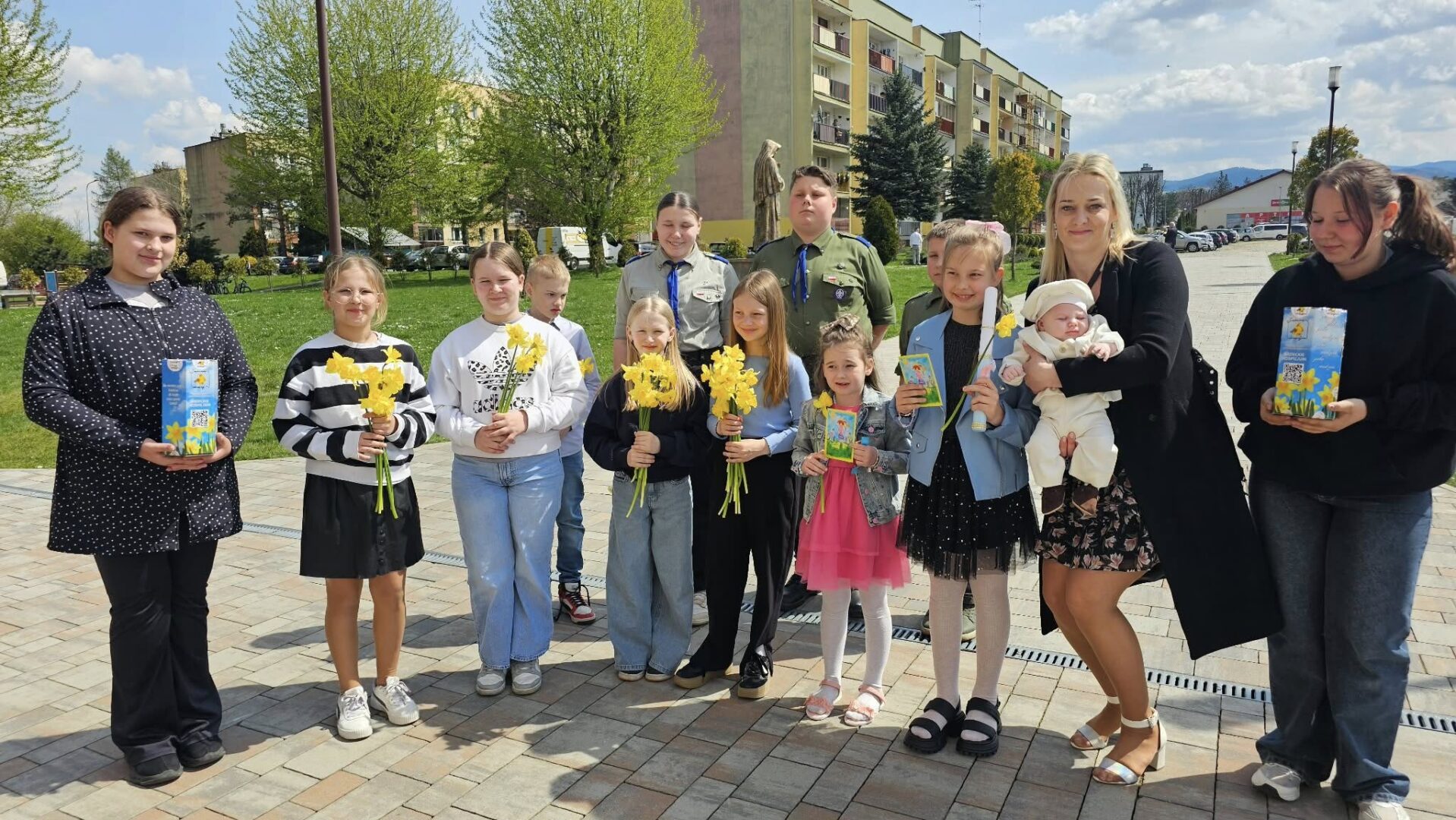 A group of children and a few adults stand outdoors in a park-like area, many holding yellow daffodils and gift cards, smiling at the camera.
