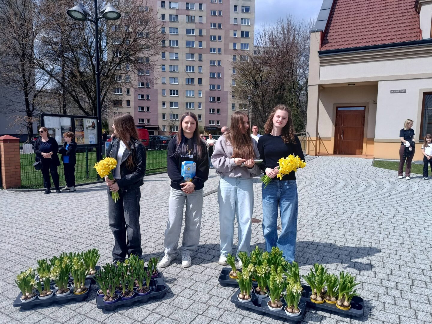 Four young women stand on a paved courtyard holding yellow bouquets, with trays of potted daffodils in front and apartment buildings behind them.
