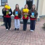 Four girls standing on a brick pathway outside a building, each holding a yellow bouquet and a blue card think?