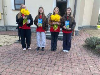 Four girls standing outdoors, each holding a bouquet of yellow flowers and a card.