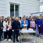 Group of people posing outdoors beside a white building with three cakes on a metal serving cart in front of them.