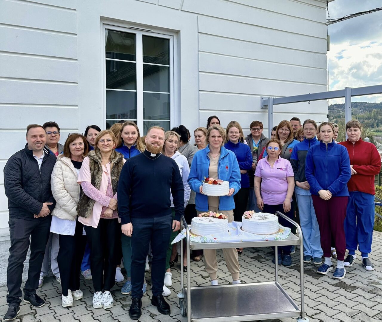 Group of people posing outdoors beside a white building with three cakes on a metal serving cart in front of them.