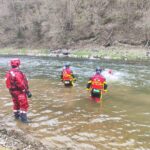 Rescue workers in red wetsuits and helmets stand knee-deep in a river, preparing to assist a swimmer further downstream.