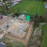 Aerial view of a construction site beside a soccer field, showing a foundation pit, concrete blocks, and a yellow excavator with a white van on site.