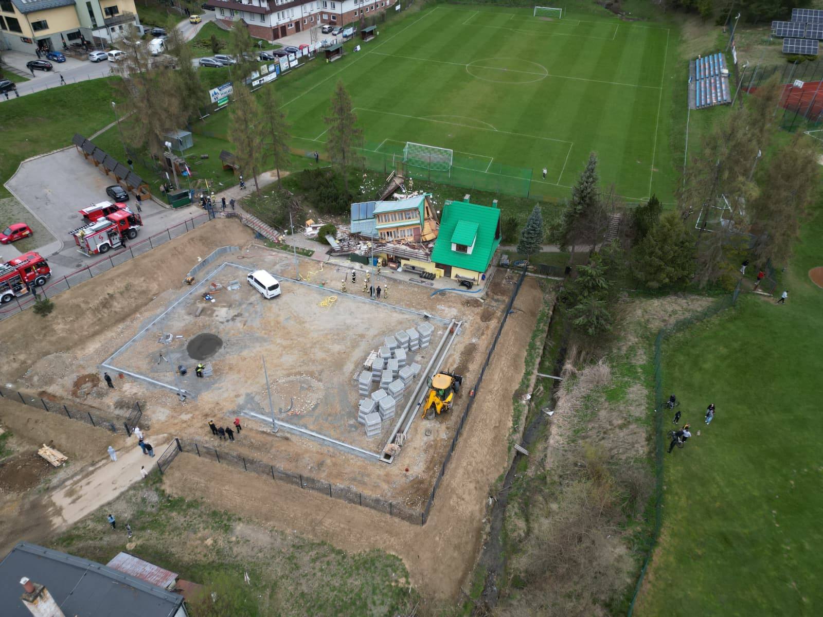 Aerial view of a construction site beside a soccer field, showing a foundation pit, concrete blocks, and a yellow excavator with a white van on site.