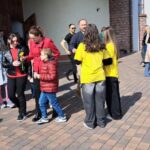 Group of people standing outside a brick building on a brick-paved area, including two people in yellow shirts talking nearby.