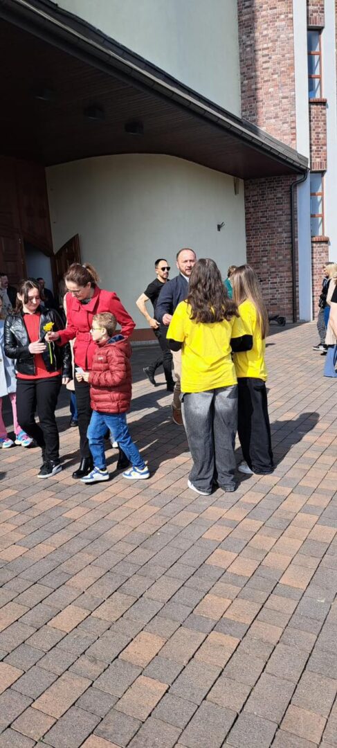 Group of people standing outside a brick building on a brick-paved area, including two people in yellow shirts talking nearby.