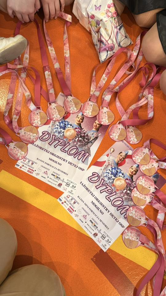 Awards laid out on an orange floor: pink-ribbon medals and diplomas labeled 'Dyplom' with dancer graphics, from a children's ceremony.