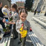 Girl in a uniform holding a bouquet of yellow daffodils at an outdoor table with other students and volunteers nearby.