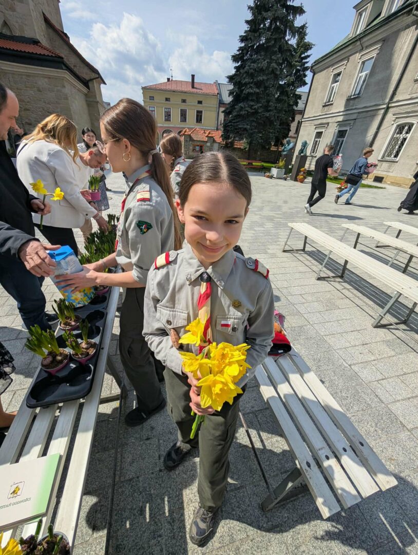 Girl in a uniform holding a bouquet of yellow daffodils at an outdoor table with other students and volunteers nearby.