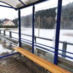 Bus stop shelter with a wooden bench on a snowy platform, blue metal frame and curved translucent roof.