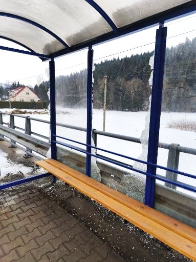 Bus stop shelter with a wooden bench on a snowy platform, blue metal frame and curved translucent roof.