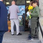Two boys in green scout uniforms hold yellow flowers and papers while standing on steps outside a doorway as adults enter a building.