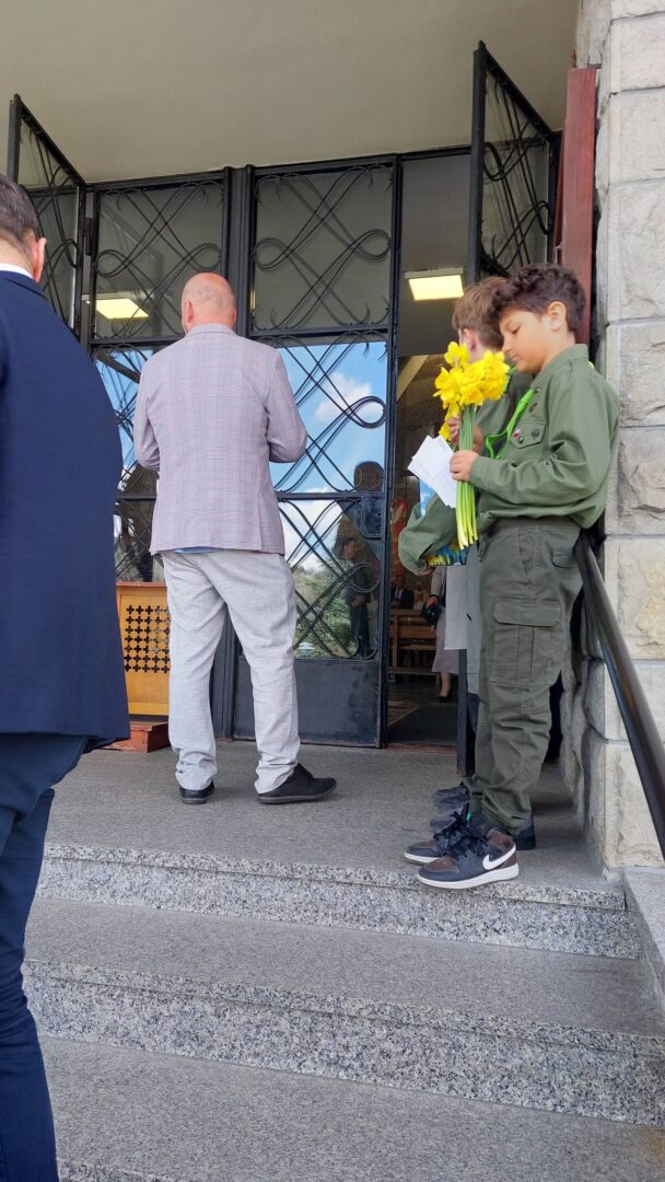 Two boys in green scout uniforms hold yellow flowers and papers while standing on steps outside a doorway as adults enter a building.