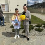 Boy in a white ceremonial robe with a gold cape, holding a booklet, standing beside a woman in a checkered suit who holds a yellow bouquet outside a church.