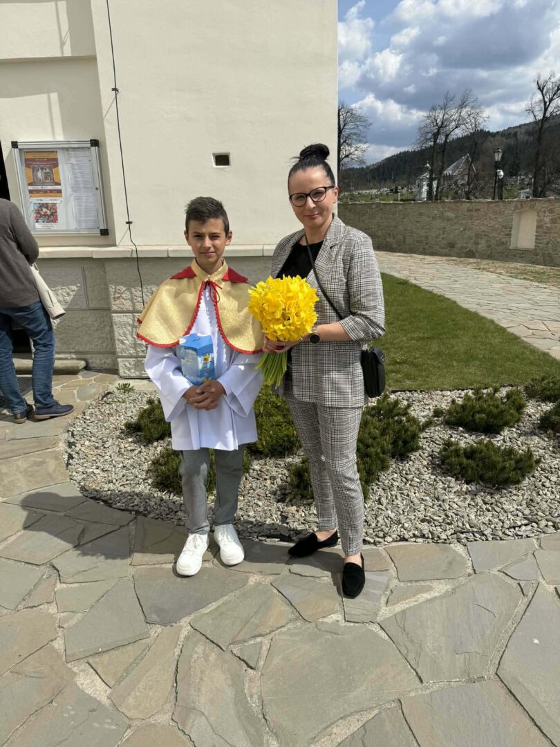 Boy in a white ceremonial robe with a gold cape, holding a booklet, standing beside a woman in a checkered suit who holds a yellow bouquet outside a church.