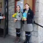 Two girl scouts in gray uniforms pose on a stone step outside a building, one holding a bouquet of yellow flowers and the other a blue book.