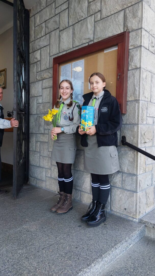 Two girl scouts in gray uniforms pose on a stone step outside a building, one holding a bouquet of yellow flowers and the other a blue book.