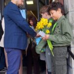 Man in a blue suit hands a card to a boy holding yellow daffodils outside a doorway; other people watch in the background.