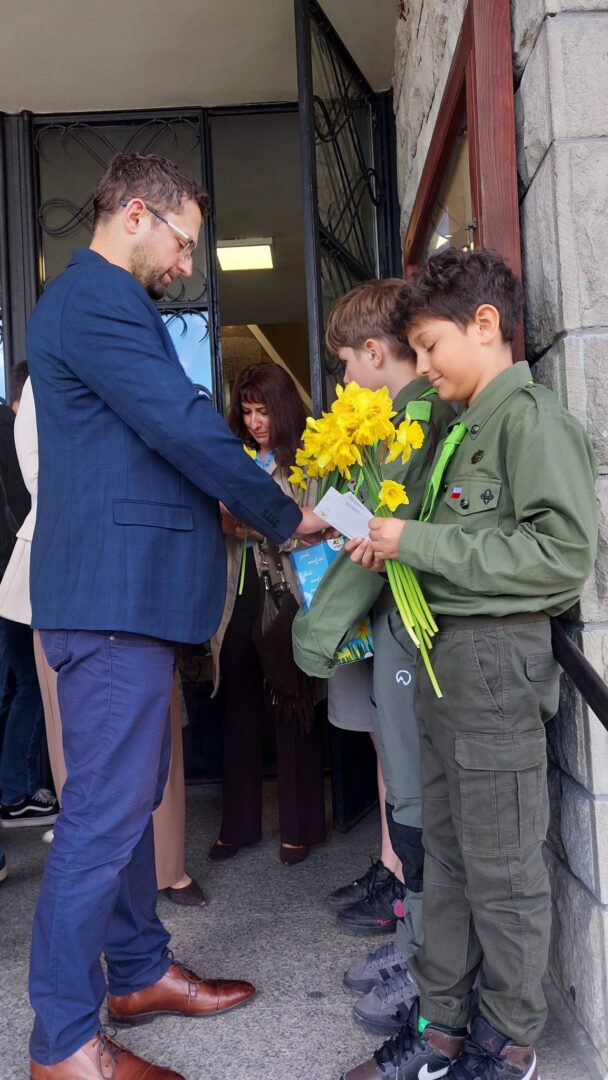 Man in a blue suit hands a card to a boy holding yellow daffodils outside a doorway; other people watch in the background.