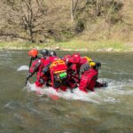 Rescue team in red wetsuits and helmets huddles together in a fast-moving river, preparing to move downstream.