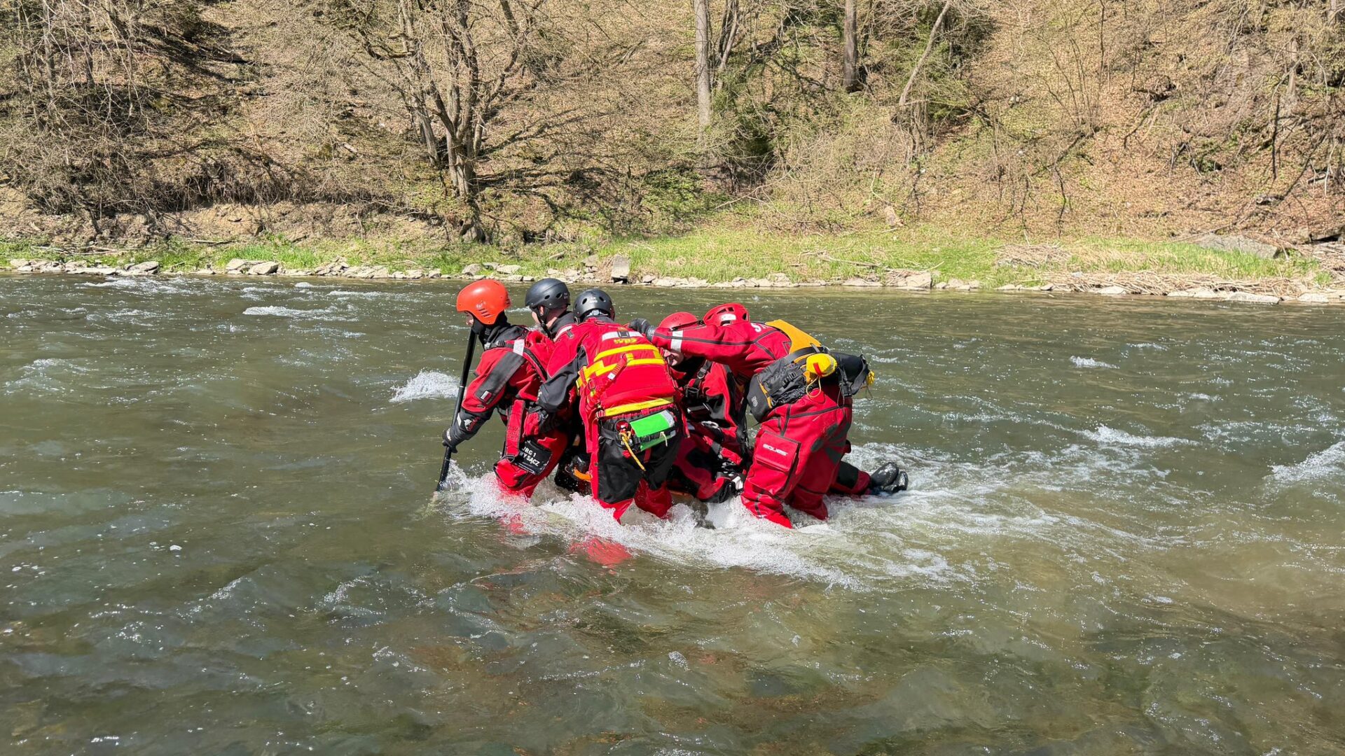 Rescuers in red wetsuits and helmets huddle in a river, using a paddle for stability during a swift current.