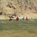 Group of people wearing life jackets and helmets wading in a shallow river near a rocky, wooded bank