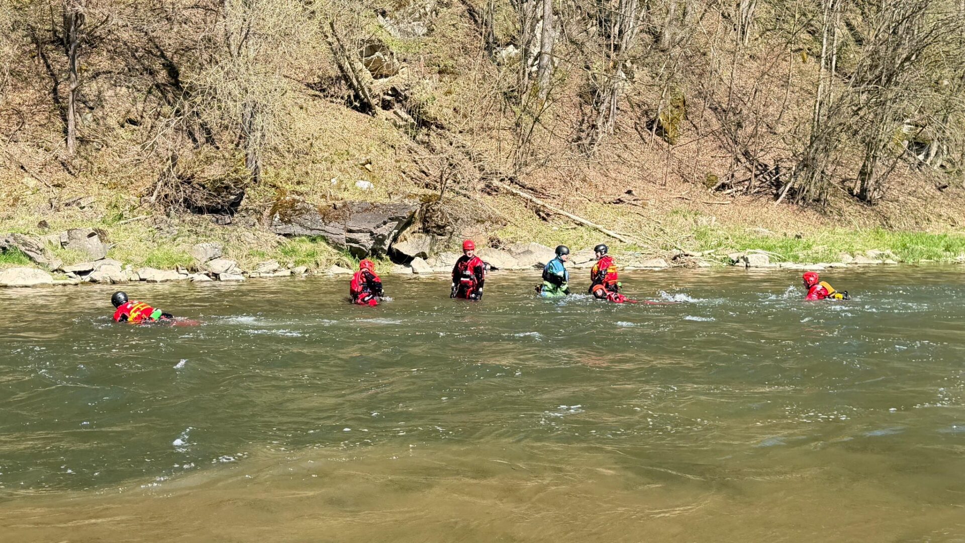 Group of people wearing life jackets and helmets wading in a shallow river near a rocky, wooded bank