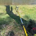 Yellow garden tool with a handle lying on grass near a riverbank; shadow of a person cast on the ground, trees across the water in the background.