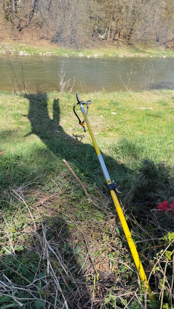 Yellow garden tool with a handle lying on grass near a riverbank; shadow of a person cast on the ground, trees across the water in the background.