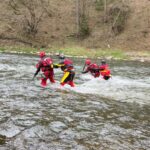 Five rescue workers in red and yellow gear and helmets cross a flowing river, assisting each other near a wooded riverbank.