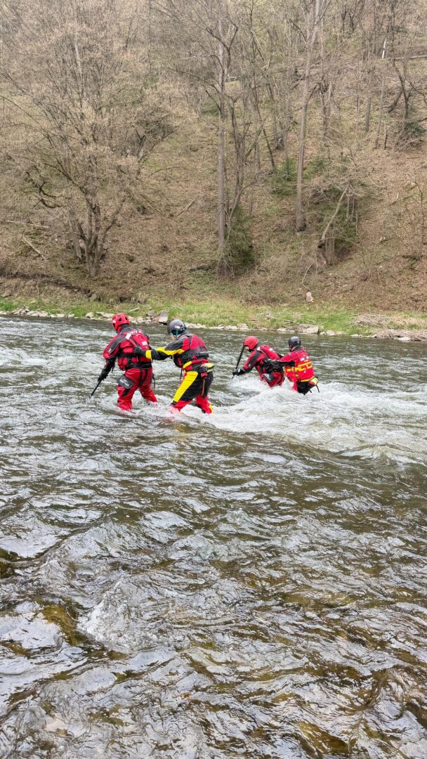 Five rescue workers in red and yellow gear and helmets cross a flowing river, assisting each other near a wooded riverbank.