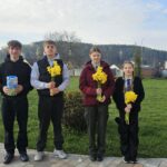 Four teenagers stand in a park at daytime, each holding a bouquet of yellow daffodils, with a statue and houses in the background.