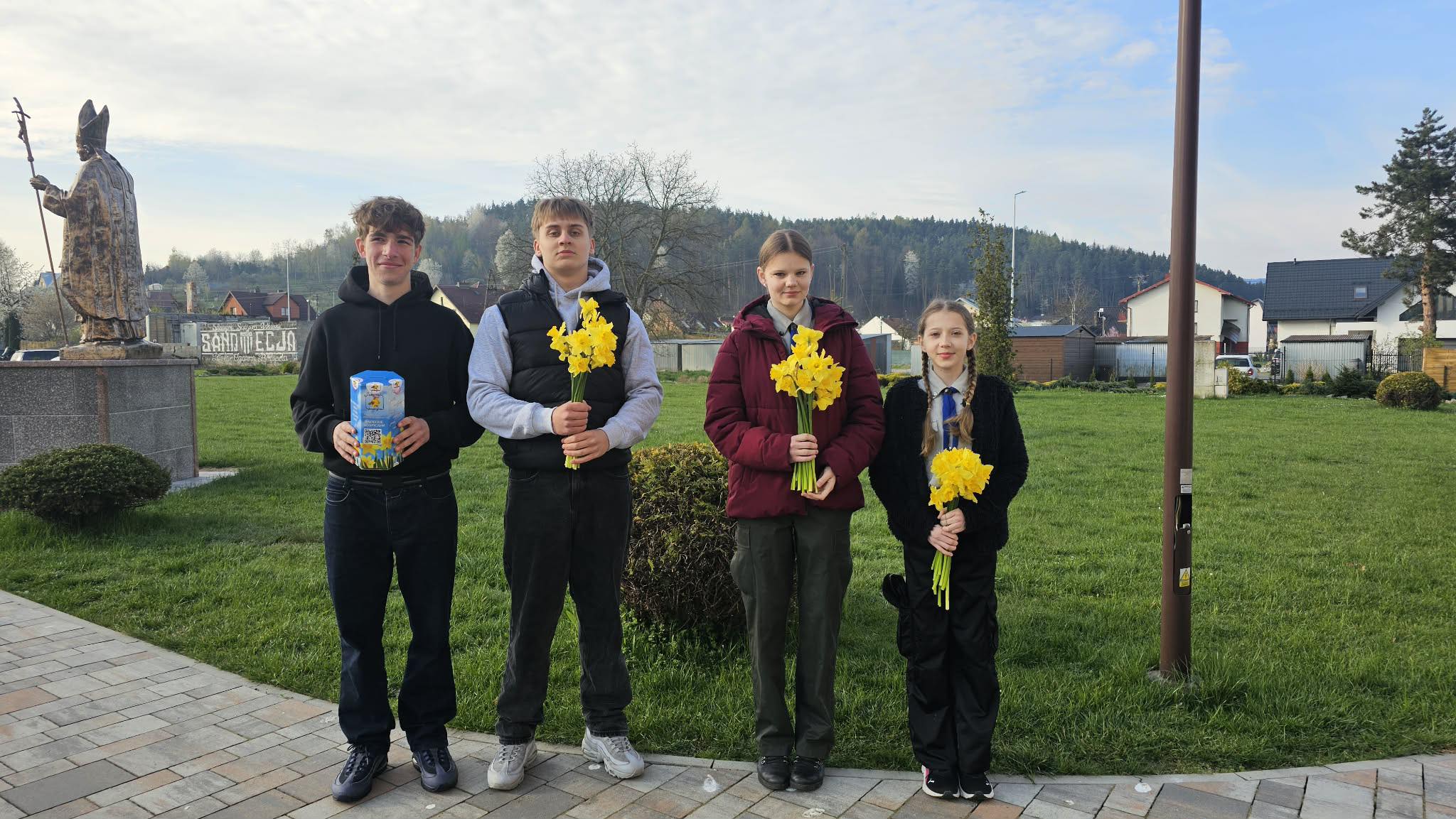 Four teenagers stand in a park at daytime, each holding a bouquet of yellow daffodils, with a statue and houses in the background.