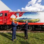 Two men in suits high-five in front of a red muraled train locomotive outdoors on a sunny day