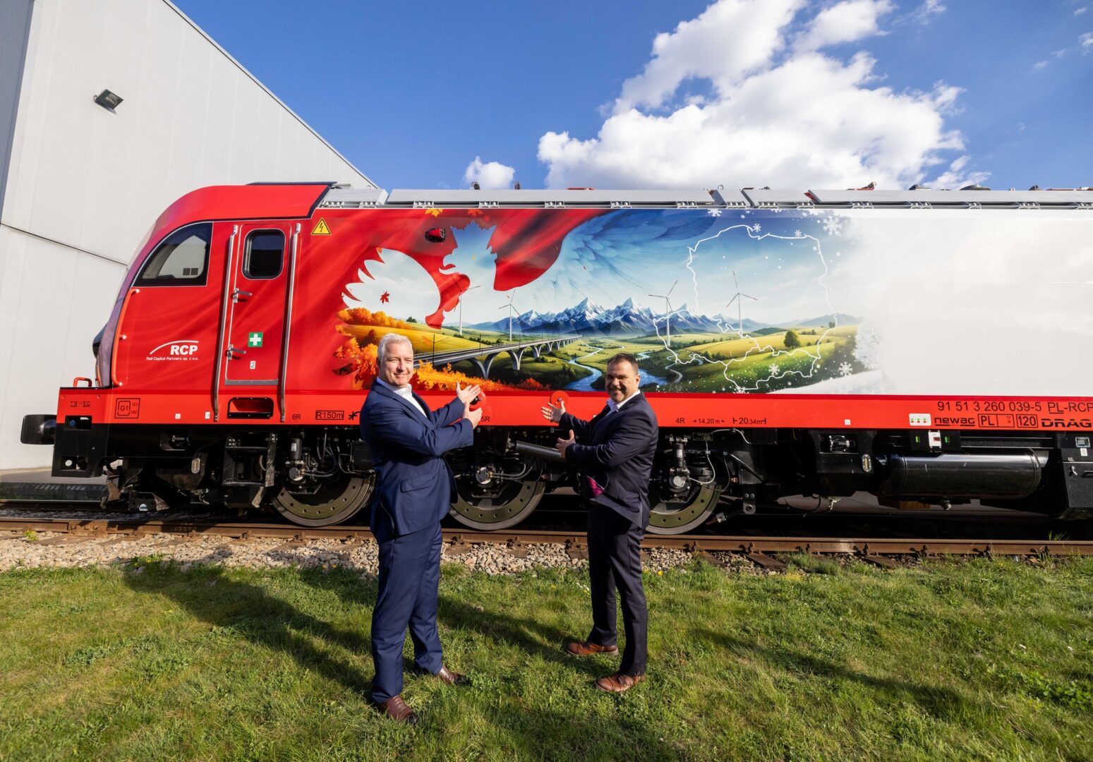 Two men in suits high-five in front of a red muraled train locomotive outdoors on a sunny day